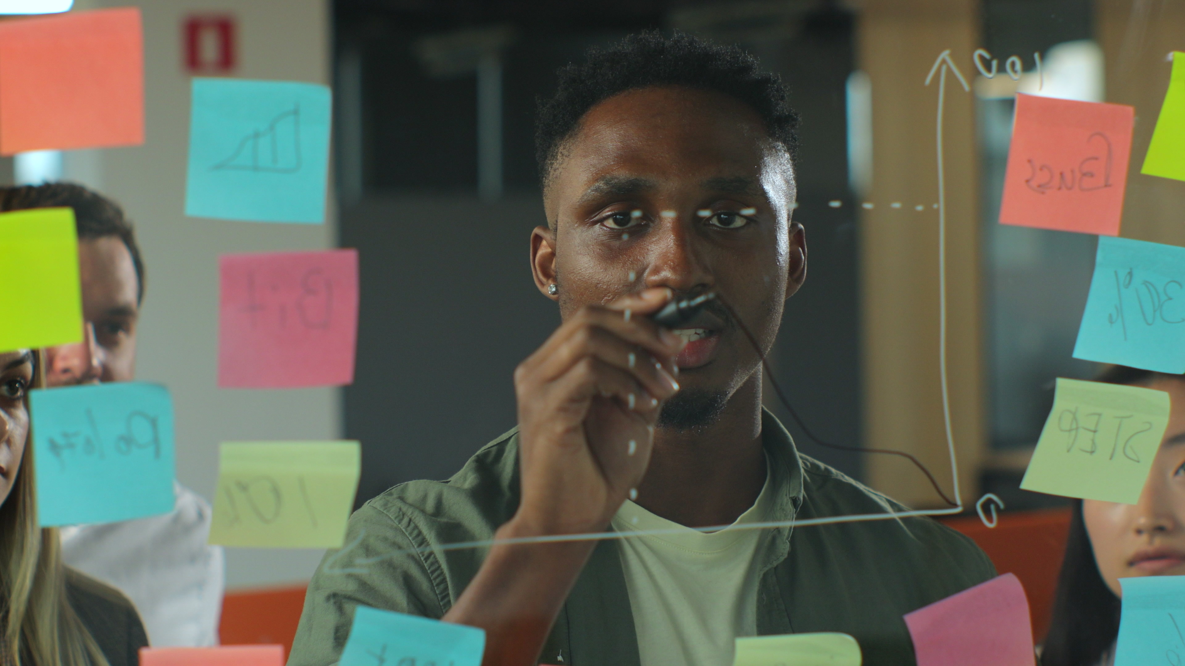 young-afro-american-man-casualwear-drawing-business-chart-glass-board-with-marker-speaking-team-coworkers-during-informal-meeting-office